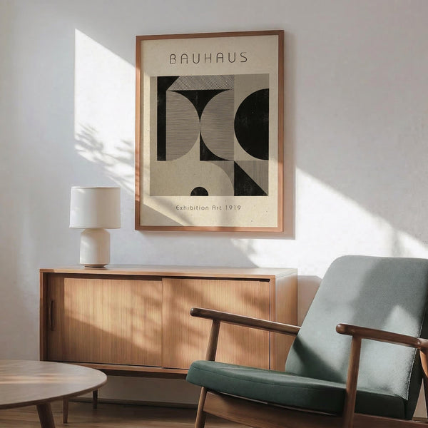 May include: Framed Bauhaus-style poster with geometric black and beige abstract design hanging above mid-century modern wooden sideboard. Scene shows mint green upholstered armchair with wooden frame, white ceramic table lamp, and round coffee table. Warm natural lighting creates shadows across white wall in minimalist room setting.