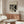 May include: Bauhaus-style poster with geometric black and beige shapes displayed above wooden sideboard in modern living room setting. White sculptural chair, glass coffee table, and minimalist table lamp complete the neutral-toned interior scene.