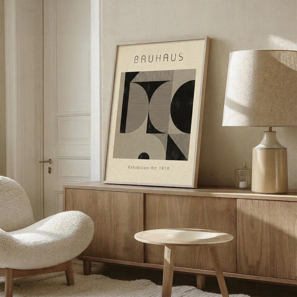 May include: Framed Bauhaus art print with geometric black and beige shapes displayed on wooden credenza in neutral living room setting. White upholstered chair, ceramic table lamp with linen shade, and small round wooden stool visible. Warm natural lighting creates soft shadows on textured beige walls.