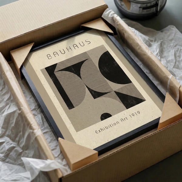 May include: Framed Bauhaus-style poster with bold geometric letter R design in black and beige, displayed in black frame inside cardboard shipping box with protective tissue paper and wooden corner guards on white surface