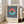 May include: Framed Bauhaus-style poster with navy and white vertical stripes, featuring a circle split into coral orange top half and turquoise bottom half. Text reads "BAUHAUS Exhibition Art 1923" below the geometric design. Light wood frame leaning against white wall next to beige sofa in bright, airy living room with large windows and hardwood floors.