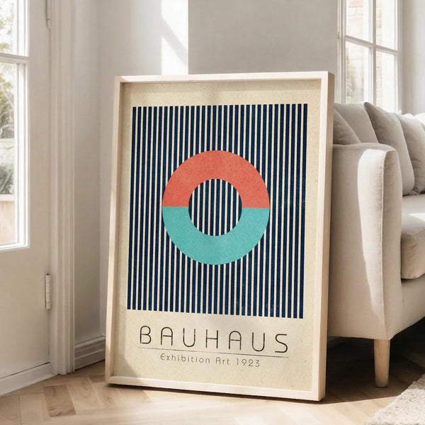 May include: Framed Bauhaus-style poster with navy and white vertical stripes, featuring a circle split into coral orange top half and turquoise bottom half. Text reads "BAUHAUS Exhibition Art 1923" below the geometric design. Light wood frame leaning against white wall next to beige sofa in bright, airy living room with large windows and hardwood floors.