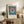 May include: Framed Bauhaus-style poster with geometric circle design in coral and teal on navy striped background, displayed on rustic wooden sideboard next to fern plant, ceramic vase, and stacked books in neutral-toned living room setting with beige armchair and warm natural lighting.