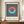 May include: Framed Bauhaus-style poster with geometric design featuring coral and turquoise half-circles forming a ring shape over navy and white vertical stripes on cream background. Wooden frame displayed on light wood sideboard next to white ceramic vase and stacked books in elegant room with white paneled walls.