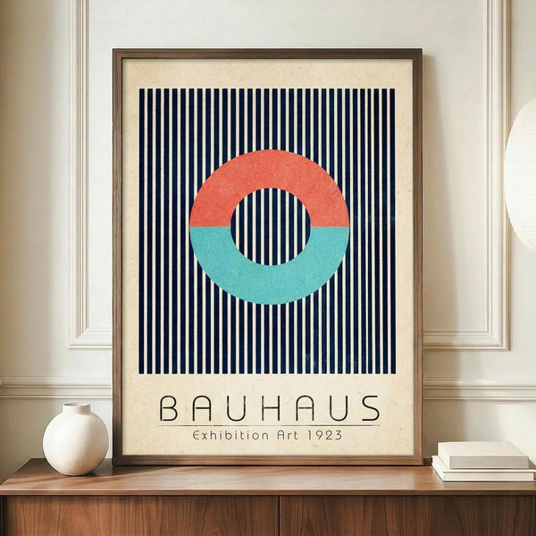 May include: Framed Bauhaus-style poster with geometric design featuring coral and turquoise half-circles forming a ring shape over navy and white vertical stripes on cream background. Wooden frame displayed on light wood sideboard next to white ceramic vase and stacked books in elegant room with white paneled walls.