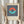 May include: Person holding framed Bauhaus-style art print featuring geometric design with coral and teal split circle over navy and cream vertical stripes, beige matting, light wood frame, dried pampas grass visible to left, neutral beige clothing and curtained background creating minimalist home decor scene