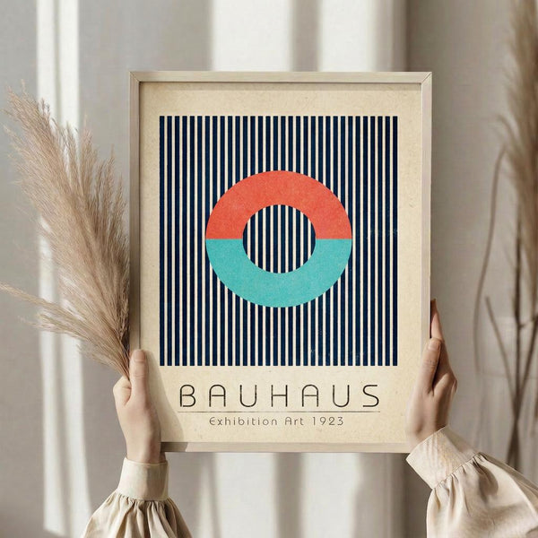 May include: Person holding framed Bauhaus-style art print featuring geometric design with coral and teal split circle over navy and cream vertical stripes, beige matting, light wood frame, dried pampas grass visible to left, neutral beige clothing and curtained background creating minimalist home decor scene