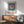 May include: Framed Bauhaus-style poster with navy blue vertical stripes, coral and teal circular design, displayed above wood sideboard in mid-century modern living room. Green upholstered armchair with wooden frame, white ceramic table lamp, and natural sunlight streaming across white wall create minimalist Scandinavian interior setting.