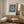 May include: Framed Bauhaus-style poster with navy vertical stripes and coral and teal semicircles forming a circle, displayed on wooden sideboard next to brass table lamp and small round wooden stool in modern neutral-toned room with cream upholstered chair and white paneled door.