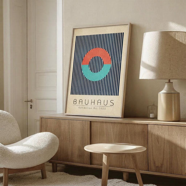 May include: Framed Bauhaus-style poster with navy vertical stripes and coral and teal semicircles forming a circle, displayed on wooden sideboard next to brass table lamp and small round wooden stool in modern neutral-toned room with cream upholstered chair and white paneled door.