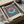 May include: Framed Bauhaus-style poster with blue and white vertical stripes background, featuring an interlocking coral and mint green circular design. Text reads "BAUHAUS Exhibition Art 1923" below the geometric pattern. The black-framed artwork sits in an open cardboard shipping box with white tissue paper, alongside a brown paper tube, on a white surface with a glass bowl visible in the background.