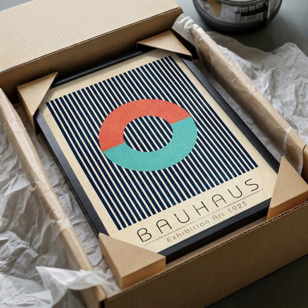 May include: Framed Bauhaus-style poster with blue and white vertical stripes background, featuring an interlocking coral and mint green circular design. Text reads "BAUHAUS Exhibition Art 1923" below the geometric pattern. The black-framed artwork sits in an open cardboard shipping box with white tissue paper, alongside a brown paper tube, on a white surface with a glass bowl visible in the background.