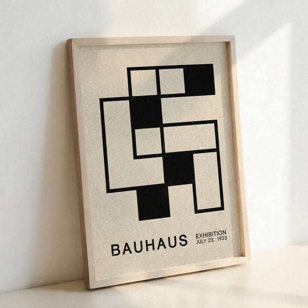 May include: Framed Bauhaus exhibition poster print leaning against white wall corner. Black geometric abstract design with interlocking rectangular shapes on cream textured background. Text reads "BAUHAUS EXHIBITION JULY 23, 1923" in black letters. Natural wood frame, minimalist modernist style artwork in bright indoor lighting.