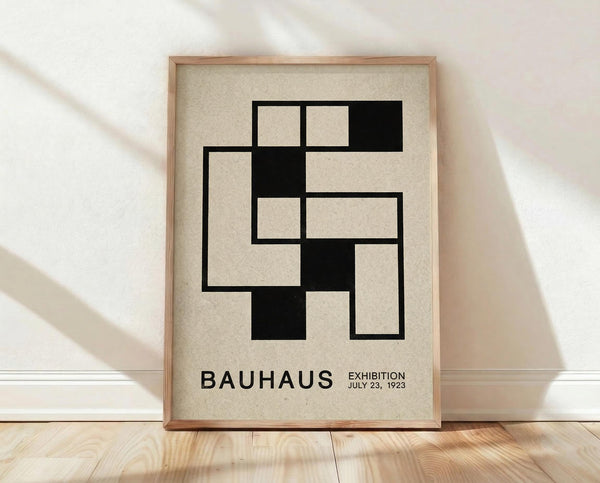 May include: Framed Bauhaus-style poster with geometric black squares forming an interlocking pattern on cream background, displayed in light wood frame against white textured wall on wooden floor with natural lighting casting diagonal shadows across the scene.