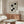 May include: Modern minimalist living room with cream walls featuring a framed Bauhaus exhibition poster with black geometric shapes on beige background. Wood credenza below holds a white dome table lamp. White curved lounge chair and glass-top oval coffee table with wood base complete the mid-century modern aesthetic. Natural lighting creates warm shadows across the neutral-toned space.