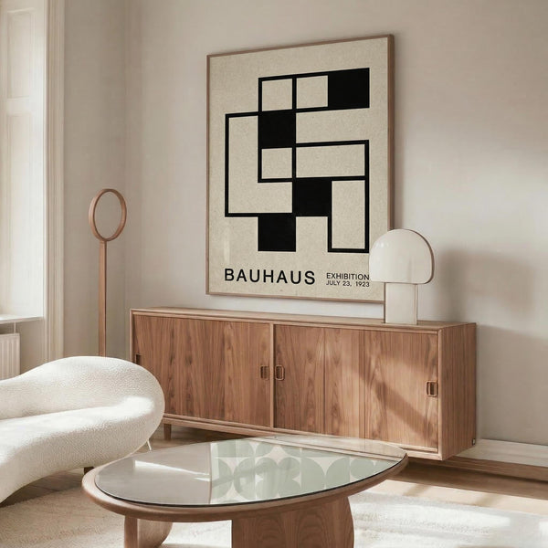 May include: Modern minimalist living room with cream walls featuring a framed Bauhaus exhibition poster with black geometric shapes on beige background. Wood credenza below holds a white dome table lamp. White curved lounge chair and glass-top oval coffee table with wood base complete the mid-century modern aesthetic. Natural lighting creates warm shadows across the neutral-toned space.
