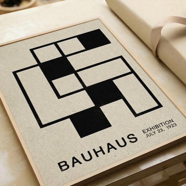 May include: Minimalist geometric art print featuring black rectangular shapes arranged in an interlocking pattern on cream background, displayed in thin wooden frame on neutral surface next to rolled paper. Modern Bauhaus-style design with "BAUHAUS EXHIBITION JULY 23, 1923" text in sans-serif font below the abstract composition.