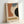 May include: Framed Bauhaus-style modernist poster leaning against white wall. Abstract geometric composition with curved organic shapes in terracotta orange, dusty blue, black, and cream on textured beige background. Text reads "JUNE 17, 1921" and "BAUHAUS" in vintage typography. Light wood frame, minimalist setting with natural lighting creating soft shadows.