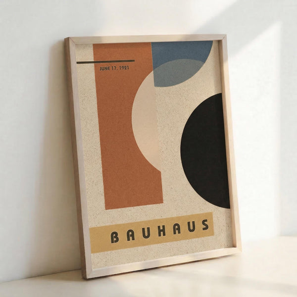 May include: Framed Bauhaus-style modernist poster leaning against white wall. Abstract geometric composition with curved organic shapes in terracotta orange, dusty blue, black, and cream on textured beige background. Text reads "JUNE 17, 1921" and "BAUHAUS" in vintage typography. Light wood frame, minimalist setting with natural lighting creating soft shadows.