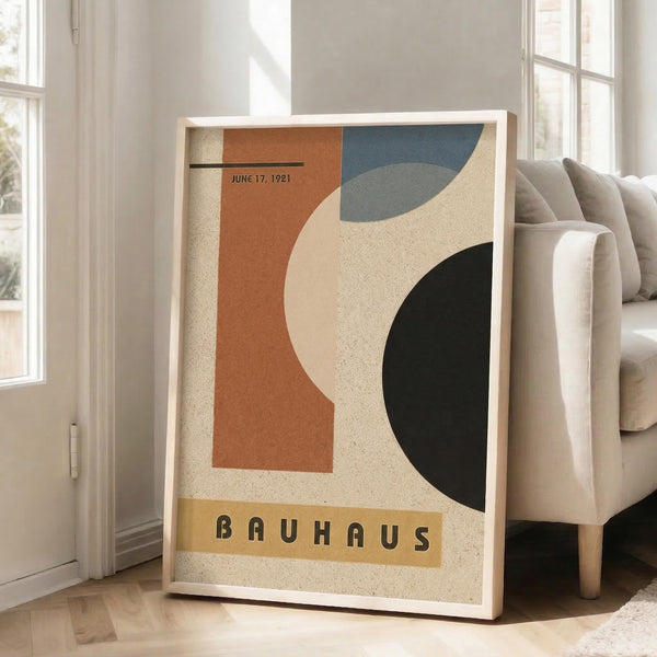 May include: Framed abstract geometric poster with orange, teal blue, cream and black curved shapes, featuring "BAUHAUS" text and "June 17, 1921" date. White frame leaning against wall next to beige upholstered chair in bright room with white curtains and hardwood floors.