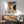 May include: Framed Bauhaus poster from June 17, 1921 featuring abstract geometric shapes in orange, blue, green, and cream colors, displayed above a mid-century modern wooden credenza with white ceramic lamp. Teal upholstered armchair with wooden frame sits nearby in bright, minimalist room with white walls and natural lighting casting dramatic shadows.