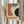 May include: Person in beige clothing holding framed abstract geometric poster featuring orange, blue, black and cream curved shapes on textured background with "BAUHAUS" text and "JUNE 17, 1921" date. Natural lighting, pampas grass visible, minimal beige interior setting.