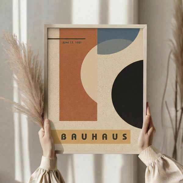 May include: Person in beige clothing holding framed abstract geometric poster featuring orange, blue, black and cream curved shapes on textured background with "BAUHAUS" text and "JUNE 17, 1921" date. Natural lighting, pampas grass visible, minimal beige interior setting.