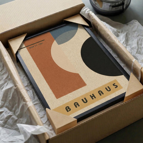 May include: Framed Bauhaus-style abstract poster with geometric shapes in terracotta, sage green, cream, and charcoal colors, featuring "JUNE 17, 1921" date marking. Black frame sits in open cardboard shipping box with white tissue paper padding. Wooden dowels for alternative hanging visible alongside, with glass vessel in background on neutral surface.