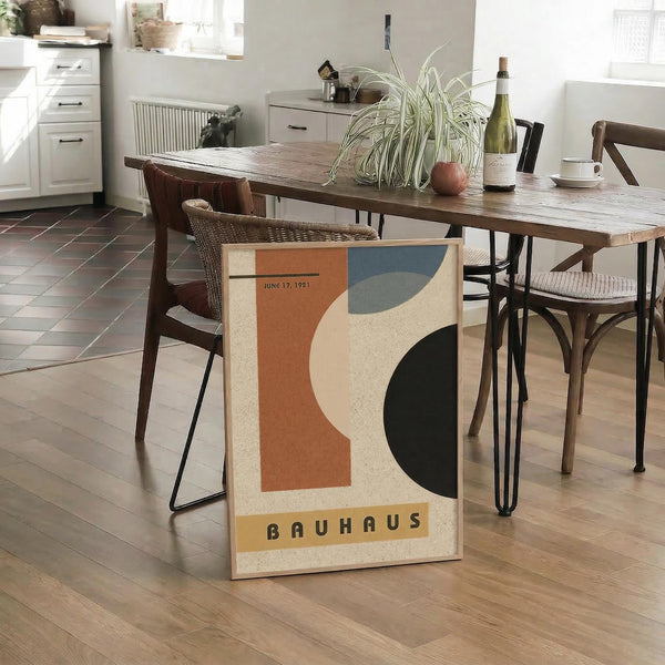 May include: Vintage Bauhaus exhibition poster dated June 17, 1921 featuring abstract geometric shapes in orange, cream, blue, and dark green colors, displayed on light wood floor against modern dining table with woven chairs in contemporary kitchen setting with white cabinets and natural lighting.