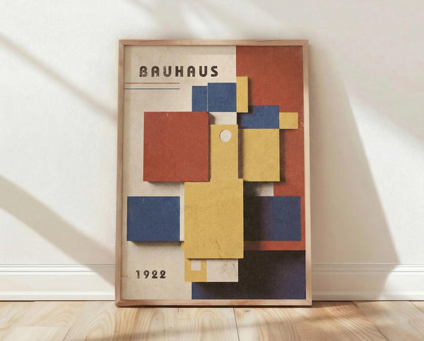 May include: Framed Bauhaus-style poster with geometric composition of overlapping colored rectangles in yellow, blue, red, and orange against cream background, displaying "BAUHAUS" and "1922" text, shown leaning against white wall on light wood floor with natural lighting casting shadows