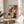 May include: Framed Bauhaus poster from 1922 featuring overlapping geometric shapes in red, blue, yellow, and white on cream background. Displayed on light wood round side table next to beige upholstered chair with plant leaves visible. Natural sunlight creates soft shadows on neutral wall. Frame appears to be thin wood finish.