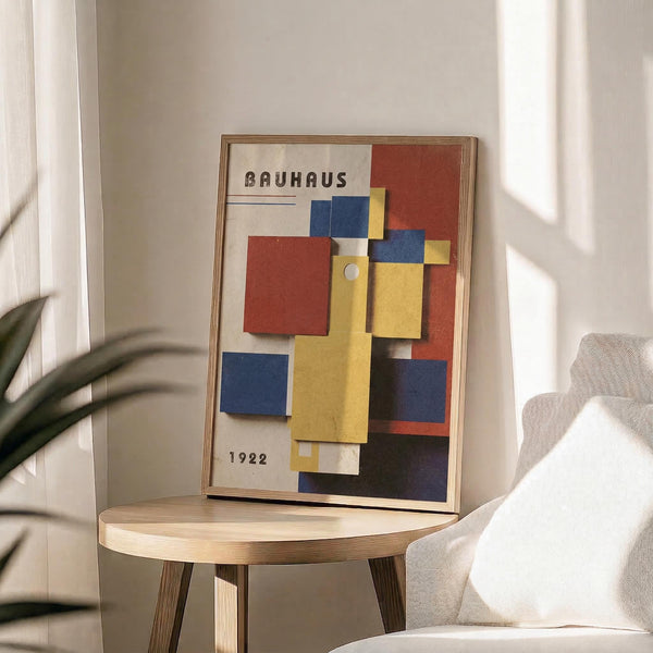 May include: Framed Bauhaus poster from 1922 featuring overlapping geometric shapes in red, blue, yellow, and white on cream background. Displayed on light wood round side table next to beige upholstered chair with plant leaves visible. Natural sunlight creates soft shadows on neutral wall. Frame appears to be thin wood finish.