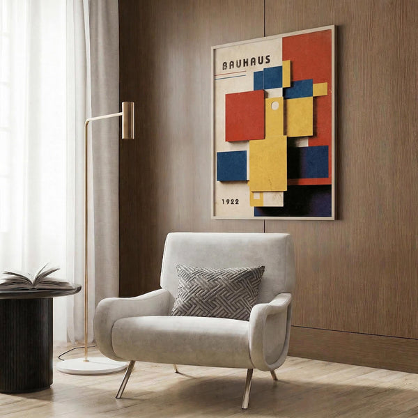 May include: Framed Bauhaus poster with colorful geometric shapes in red, yellow, blue, and orange displayed on wood paneled wall above beige upholstered armchair with patterned pillow. Modern brass floor lamp and side table with open book complete the minimalist living room scene with neutral tones and natural lighting.