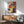 May include: Framed Bauhaus poster from 1922 featuring geometric shapes in red, blue, yellow, and white arranged in an abstract composition, displayed above a wooden mid-century credenza with white ceramic lamp, next to a sage green upholstered armchair with wooden frame in a sunlit modern living room