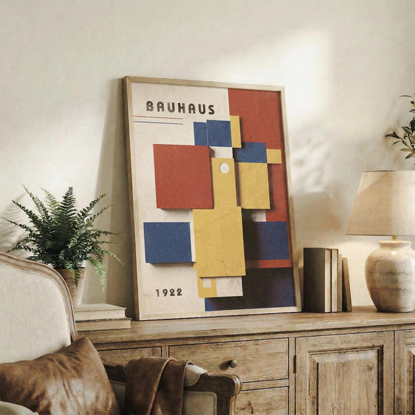 May include: Framed Bauhaus-style poster with geometric colorful rectangles in red, blue, yellow, and orange displayed on rustic wooden sideboard. Room scene shows neutral armchair, potted fern, ceramic vase, and books creating cozy living space with warm lighting and textured wall background.