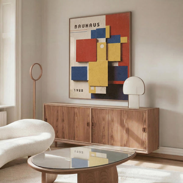 May include: Framed Bauhaus exhibition poster from 1922 featuring geometric shapes in red, blue, and yellow colors displayed above a walnut wood credenza in a modern living room. White sculptural chair and glass coffee table visible in foreground, with warm natural lighting creating reflections on surfaces.