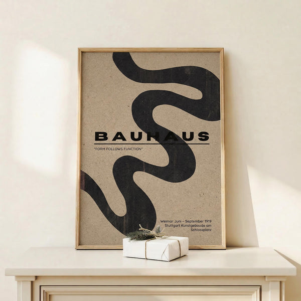 May include: Framed Bauhaus poster with black curved ribbons on beige textured background, displayed on light wooden shelf against white wall. Small white wrapped gift with twine sits nearby. Warm natural lighting creates soft shadows. Modern minimalist home decor styling with clean lines and neutral tones.