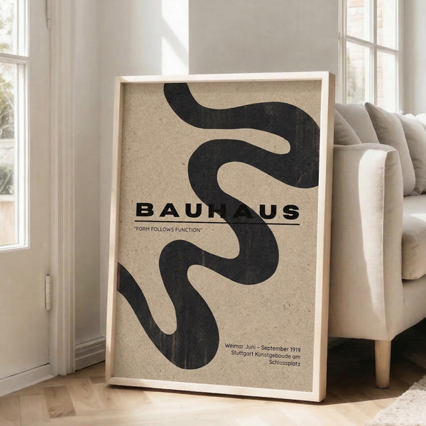 May include: A framed Bauhaus poster with flowing black curved shapes on beige textured background, featuring "Form Follows Function" text and exhibition details from Weimar 1919. The light wood frame leans against white walls next to a beige upholstered sofa in a bright, minimalist living room with large windows.