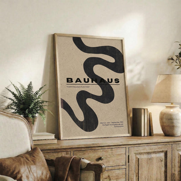 May include: Framed Bauhaus poster with flowing black abstract shape on beige background, displayed on rustic wooden sideboard beside fern plant, books, and ceramic vases in warm-toned living room setting with leather armchair