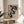 May include: Framed Bauhaus poster with black serpentine design on beige background, displaying "Form Follows Function" text and German exhibition details. Shows wooden frame resting on light wood round stool beside beige upholstered chair, with plant leaves visible and natural window light casting geometric shadows on white wall.