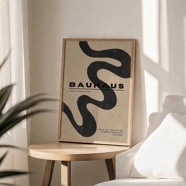 May include: Framed Bauhaus poster with black serpentine design on beige background, displaying "Form Follows Function" text and German exhibition details. Shows wooden frame resting on light wood round stool beside beige upholstered chair, with plant leaves visible and natural window light casting geometric shadows on white wall.