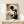 May include: Framed Bauhaus-style geometric abstract poster with black circle, rectangles, and vertical lines on cream and tan background, displayed on light wood console table beside white wrapped gift with evergreen sprig against cream wall