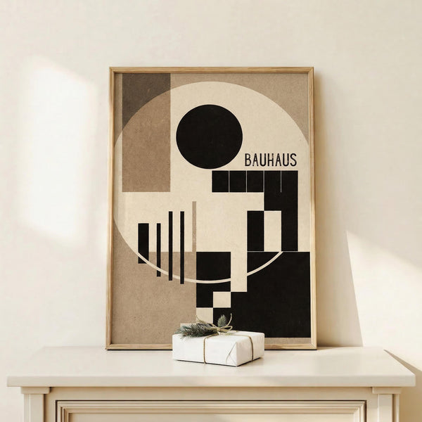 May include: Framed Bauhaus-style geometric abstract poster with black circle, rectangles, and vertical lines on cream and tan background, displayed on light wood console table beside white wrapped gift with evergreen sprig against cream wall