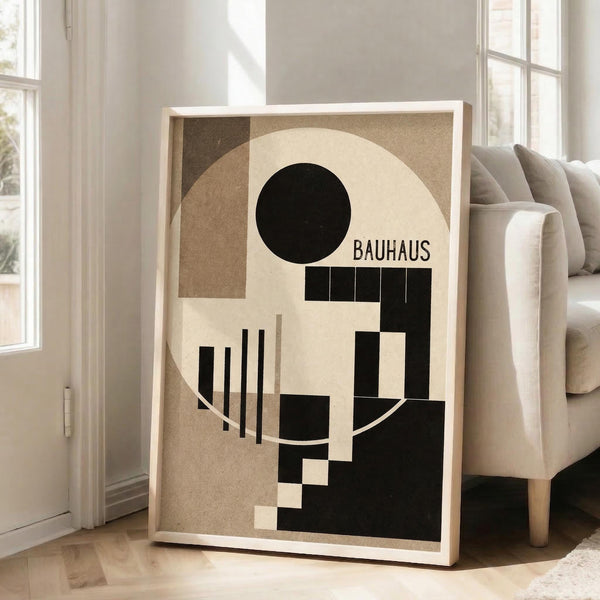 May include: Framed Bauhaus-style geometric abstract poster with black circle, beige and brown rectangles, and vertical lines against cream background, displayed in light wooden frame leaning against white wall next to beige upholstered chair in bright, airy room with large windows