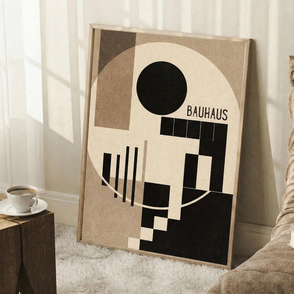 May include: A framed geometric abstract poster in earthy tones featuring black, cream, and brown shapes including circles, rectangles, and angular forms with "BAUHAUS" text. The artwork leans against a white wall next to a beige armchair, with a wooden side table holding a white coffee cup and saucer. Natural window light creates soft shadows on the white textured rug below.
