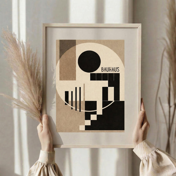 May include: Person holding framed geometric abstract poster featuring black circle, brown and cream rectangular shapes, and "BAUHAUS" text in modernist style. Light wood frame with white matting. Dried pampas grass held in other hand. Neutral beige clothing and soft natural lighting by window with white curtains create minimalist aesthetic.