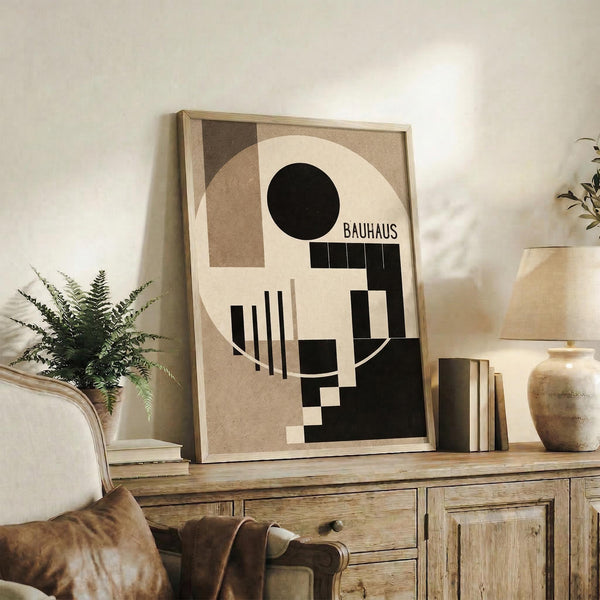 May include: Framed Bauhaus-style geometric abstract poster with black circle, beige and brown angular shapes, and vertical lines displayed on rustic wooden sideboard next to leather chair, potted fern, ceramic vases and stacked books in warm neutral living room setting.