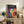 May include: Bauhaus-inspired poster with overlapping colorful circles in vibrant blues, purples, oranges, yellows, and reds on dark background, displayed in light wood frame on rustic wooden sideboard alongside fern plant, books, ceramic vases, and leather chair in warm, naturally-lit living room setting.