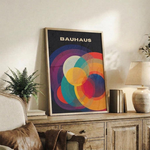 May include: Bauhaus-inspired poster with overlapping colorful circles in vibrant blues, purples, oranges, yellows, and reds on dark background, displayed in light wood frame on rustic wooden sideboard alongside fern plant, books, ceramic vases, and leather chair in warm, naturally-lit living room setting.