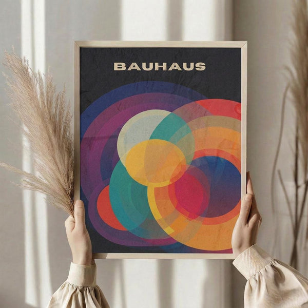 May include: Person holding framed Bauhaus-style poster with overlapping translucent circles in vibrant colors including blue, purple, red, orange, yellow, and teal against black background. Light wood frame, dried pampas grass visible, neutral beige clothing and curtains in soft natural lighting.